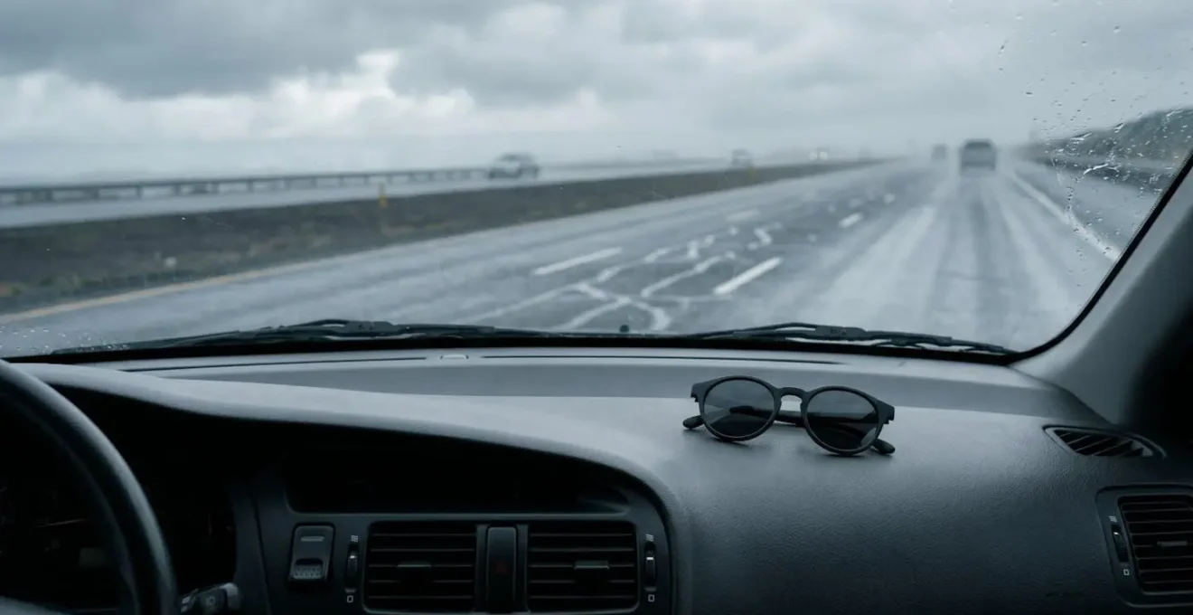 Gafas de sol polarizadas sobre salpicadero de coche con parabrisas reflejando carretera mojada en día nublado